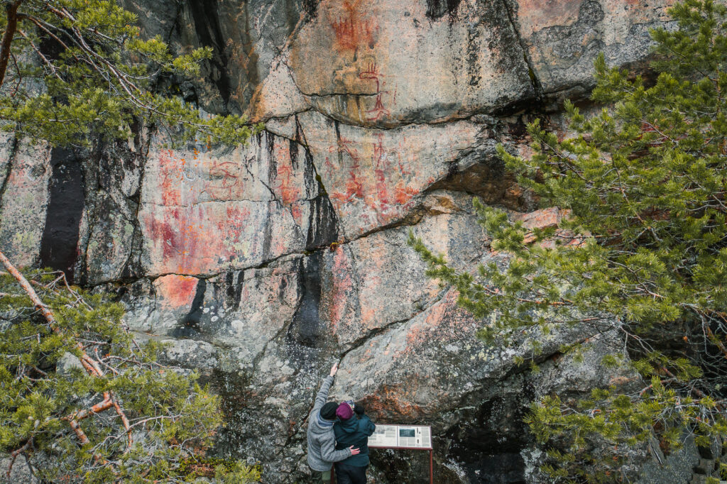 Kuvassa Astuvansalmen kalliomaalauksia. Kalliomaalaukset näkyvät punaisella värillä kallioseinämässä. Kalliota ympäröi mäntyjä. Kuvan alareunassa keskellä pariskunta ihastelee kalliomaalauksia. 
