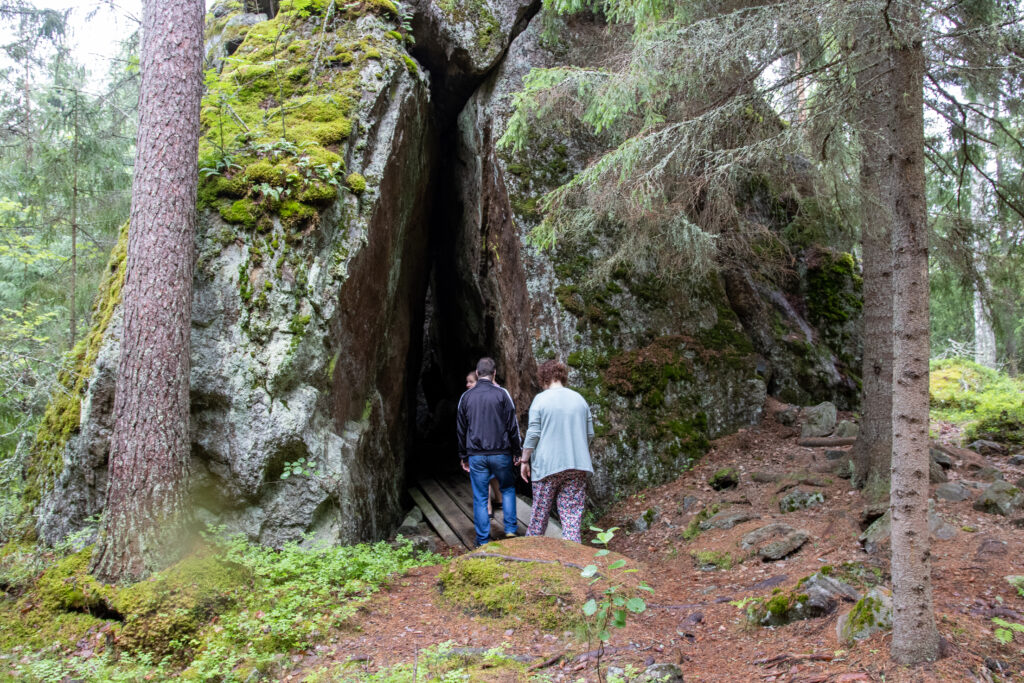 Kuvassa Karjulanmäen kivikirkko, minkä uumeniin kulkemassa kolme ihmistä metsän keskellä.