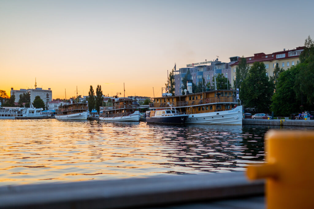 Kuva Savonlinnan matkailusatamasta ilta-auringossa. Edustalla vesistöä. Laiturissa on kolme vanhaa höyrylaivaa odottamassa risteilijöitä. 